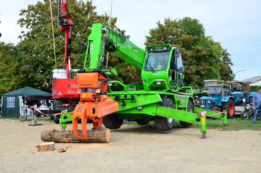 Grüner Teleskopstapler auf ausgefahrenen Stützen, daneben ein roter LKW mit Ladekran.